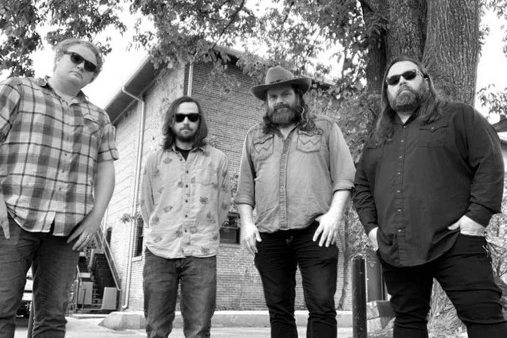 A modern photo in black and white of four men in sunglasses stand outside under a tree