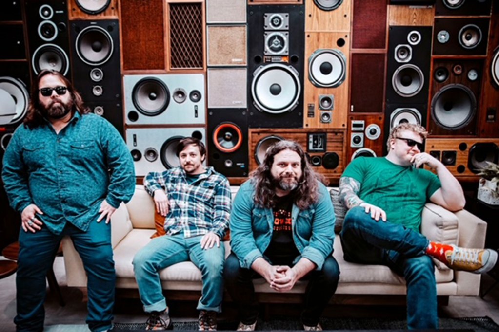 Four men sit on a couch in front of. decorative wall of music speakers
