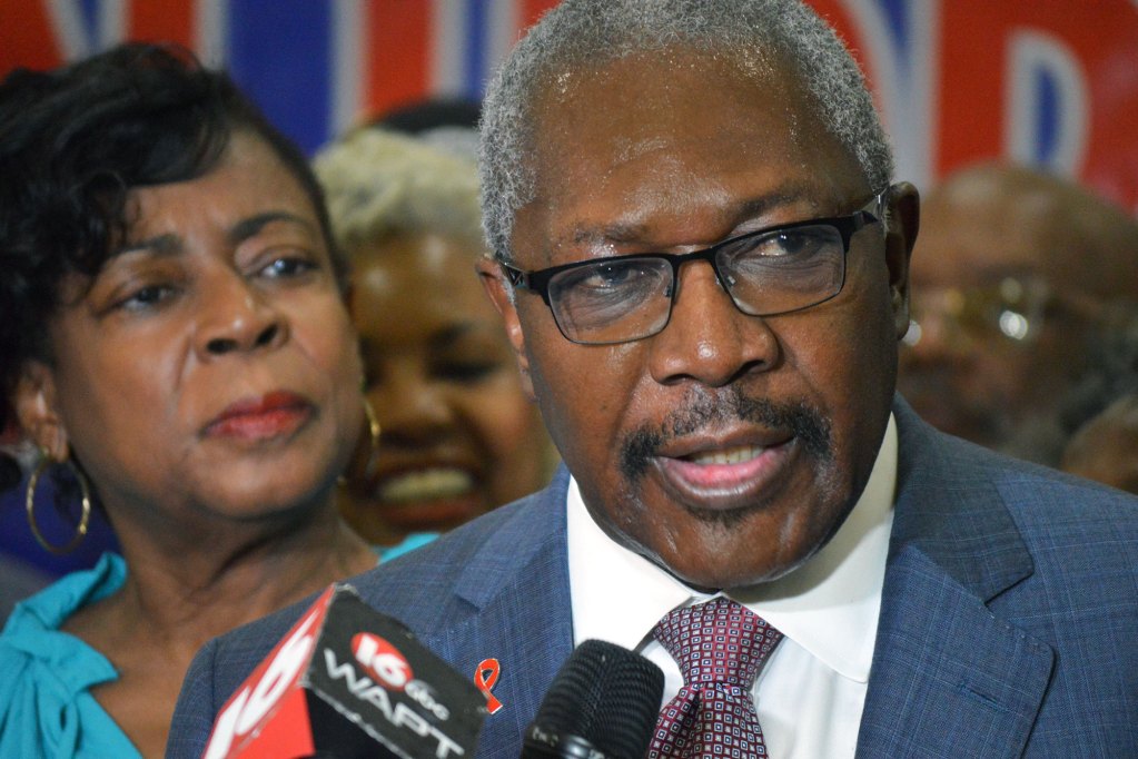 A man in glasses and a suit speaks int a reporter's mic as his wife and crowd look on behind him