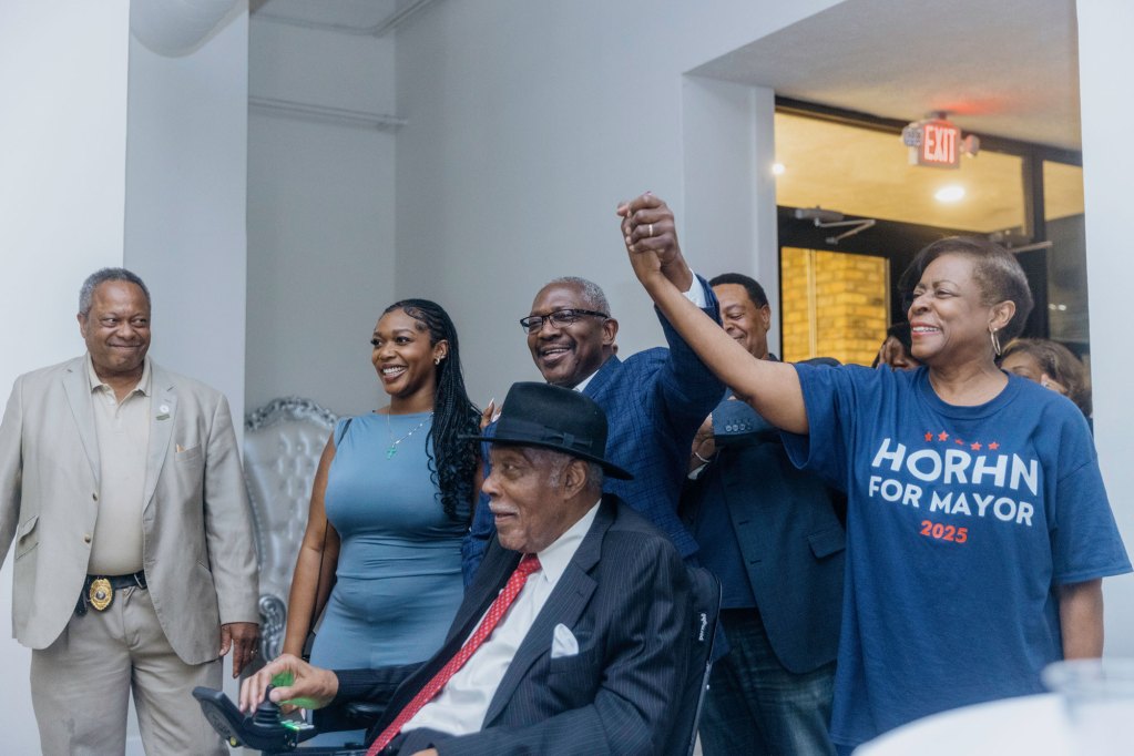 John Horhn and family enters election party wearing blue and red shirts, his wife holding up his hand while his daughter stands at his side and his father, an older man, enters in a wheelchair. Everyone is smiling