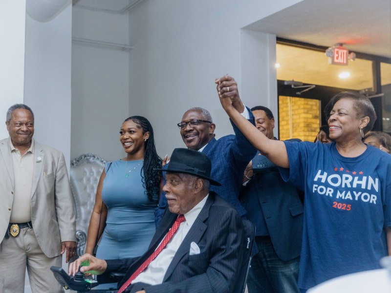 John Horhn and family enters election party wearing blue and red shirts, his wife holding up his hand while his daughter stands at his side and his father, an older man, enters in a wheelchair. Everyone is smiling
