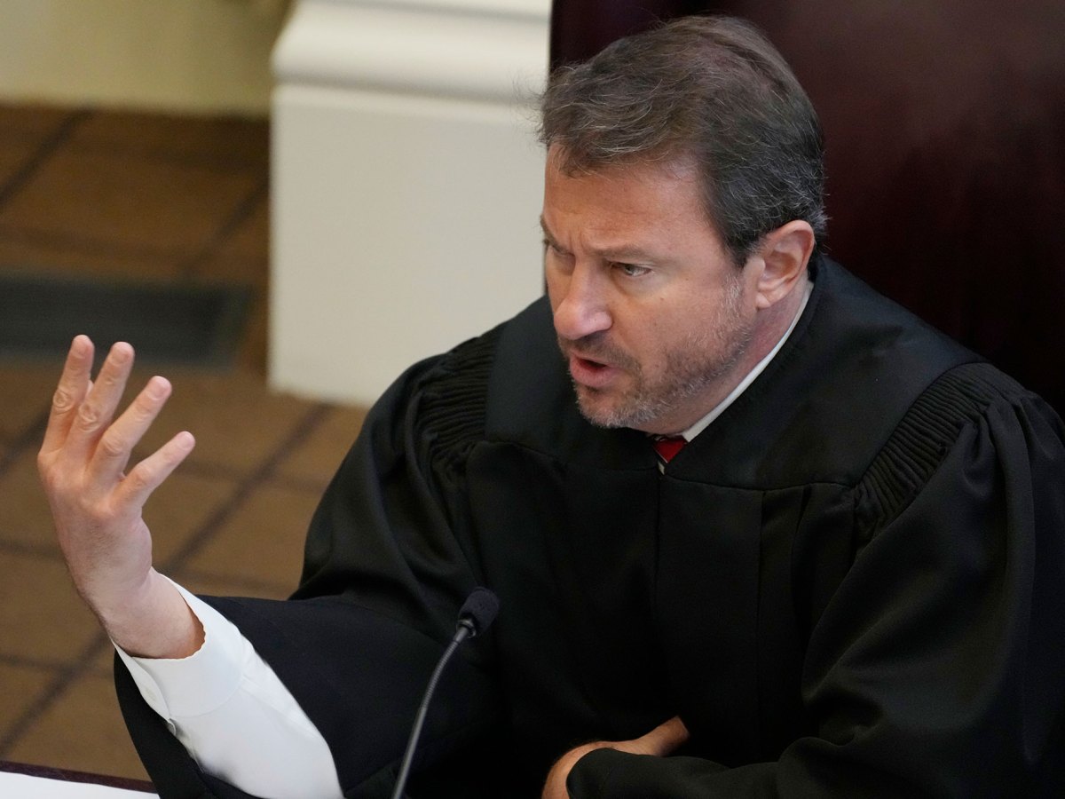 A judge, in black robes, gestures with one hand while speaking from the bench