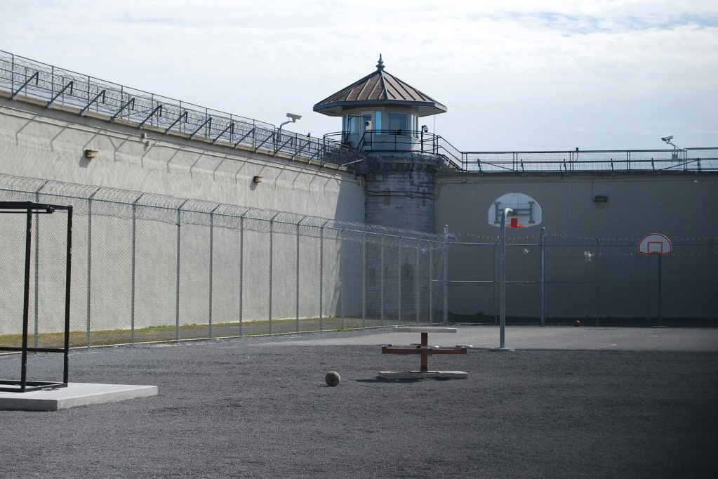 A prison yard with a fence and barbwire and two basketball goals
