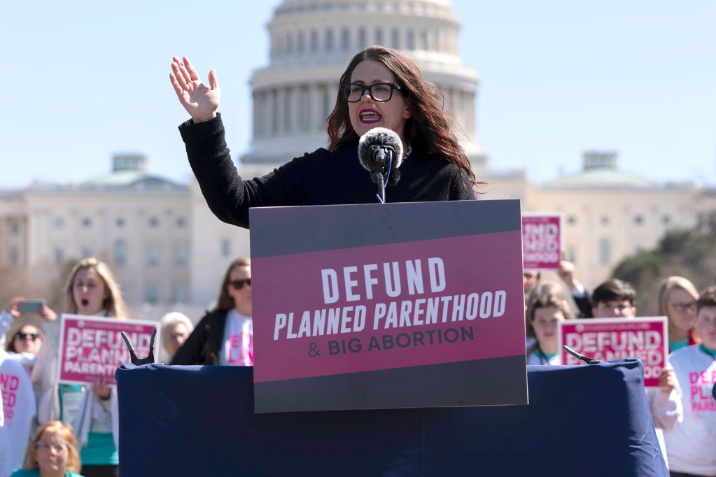 a woman speaking in front of the U.S. Capitol in front of a sign that says “Defund Planned Parenthood”