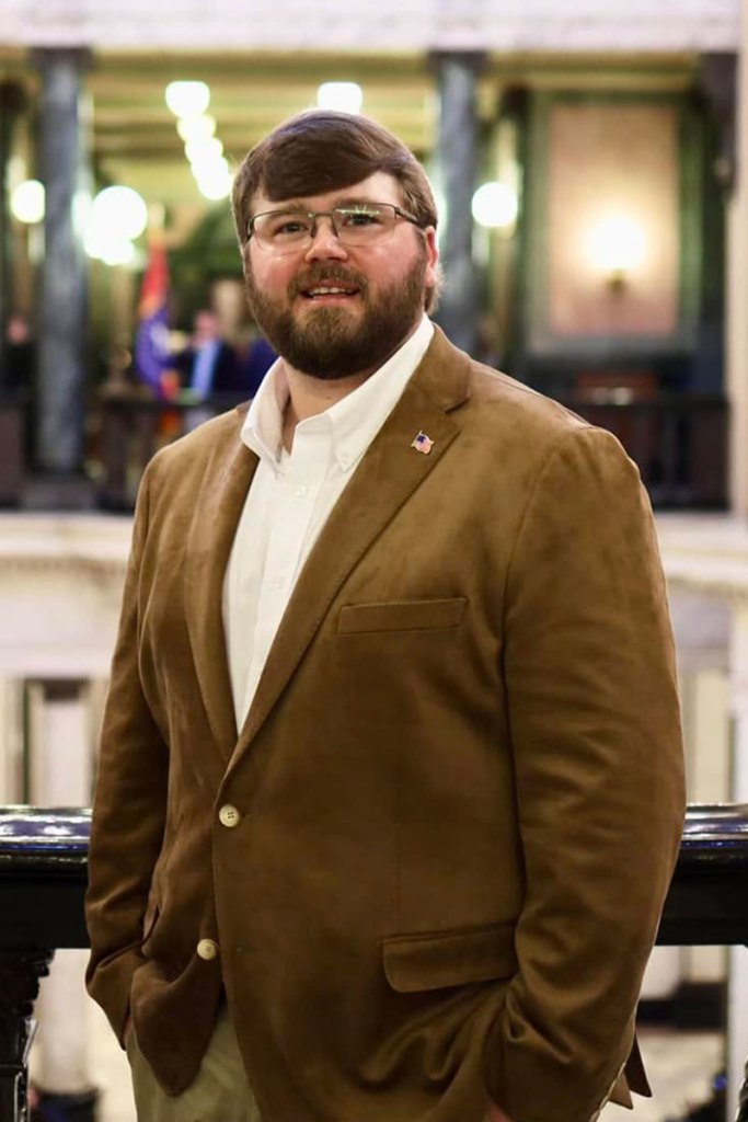 A man wearing a brown jacket poses inside the capital building