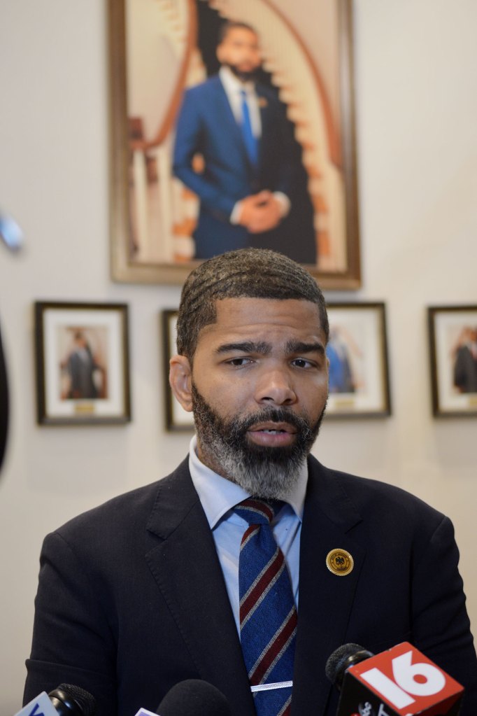 Mayor Chokwe A. Lumumba speaking in a mic wearing a navy blazer with a blue, red, and grey tie with his official portrait behind him.