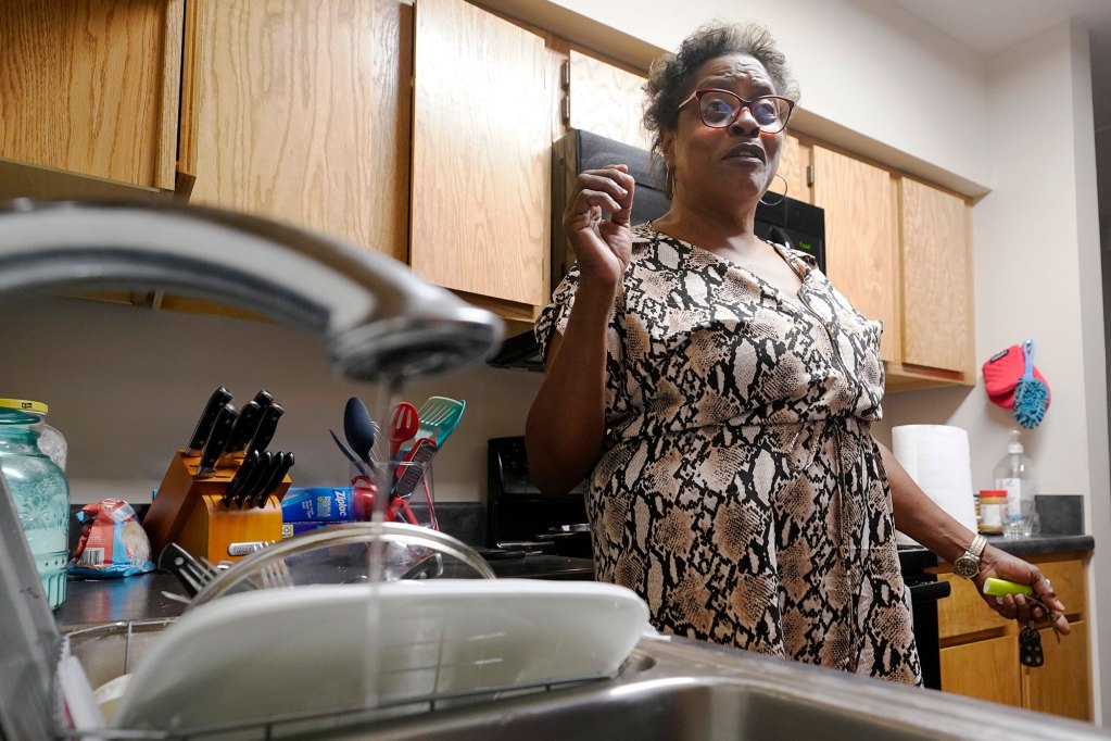A woman stands in her kitchen as a sink in the foreground shows a trickle of water
