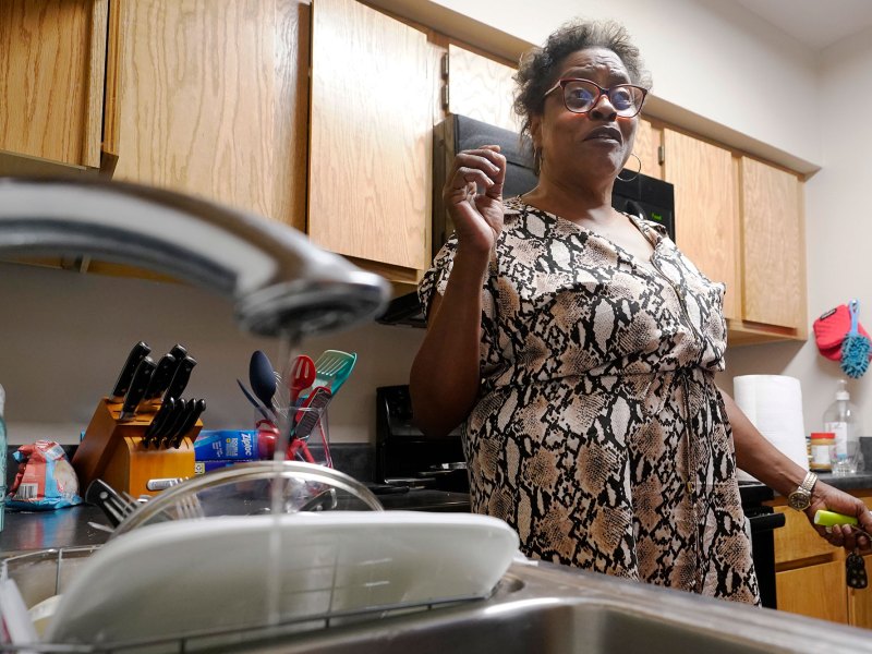 A woman stands in her kitchen as a sink in the foreground shows a trickle of water