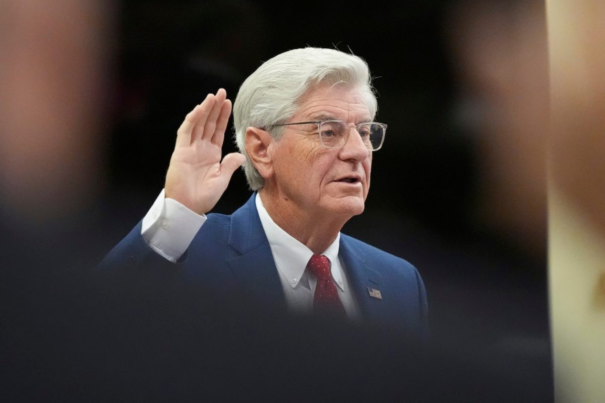 a photo showing an older man with glasses and coiffed hair, raising his hand