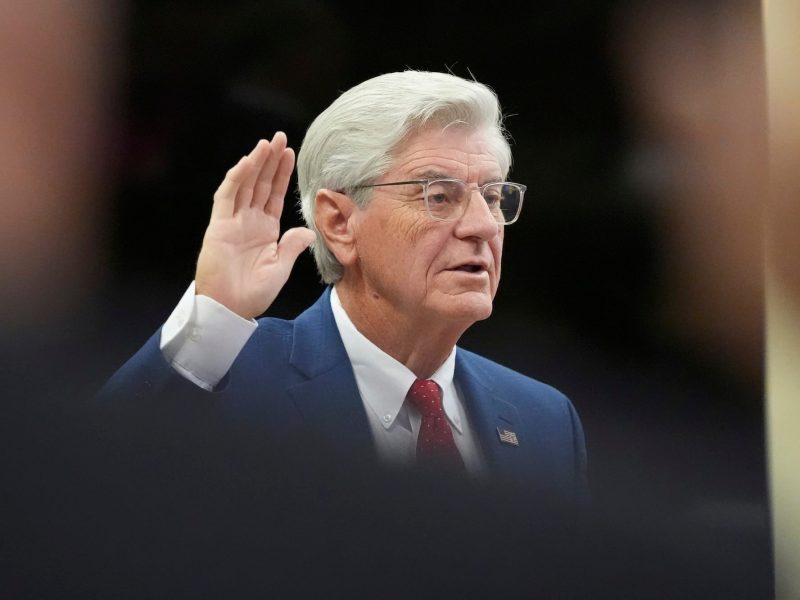 a photo showing an older man with glasses and coiffed hair, raising his hand