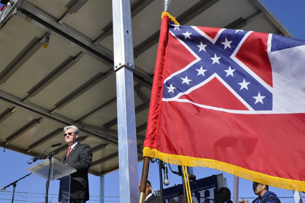 An old white man speaking at a lectern while a flag containing the Confederate battle emblem and a red, white and blue stripe flies beside him