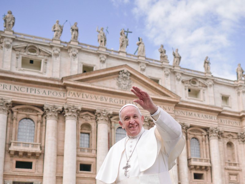 Pope Francis, in white, smiles and waves from in front of a Vatican building