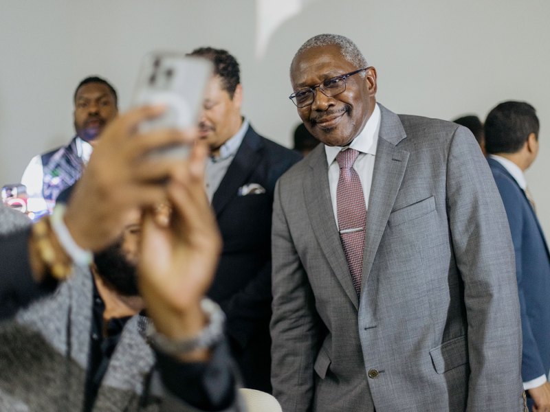John Hohrn in a suit watching as someone's arm is reached out holding a cell phone to take a photo