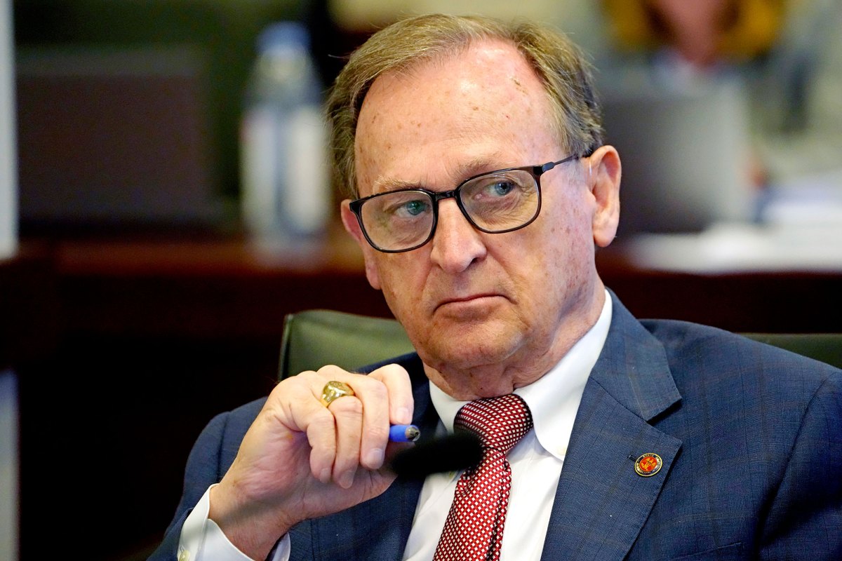 Senator Dean Kirby wearing a blue stripped suit Jacket with a red tie holds a blue pen in hand while looking to his left.