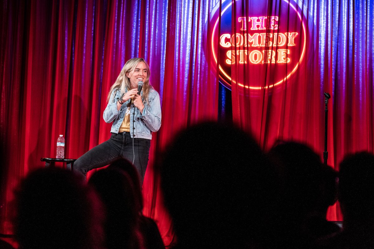 A woman sitting in front of a red velvet back drop with the Comedy Store written on it.