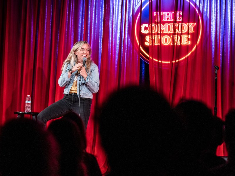 A woman sitting in front of a red velvet back drop with the Comedy Store written on it.