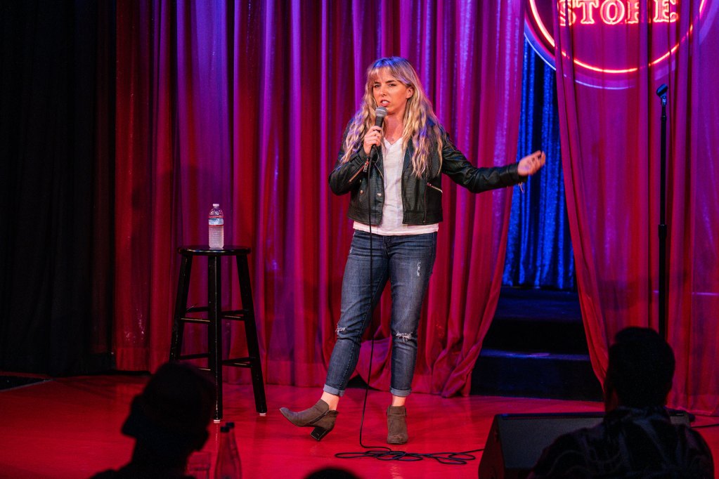 A woman speaks before a crowd as she stands in front of a red velvet back drop with the Comedy Store written on it.