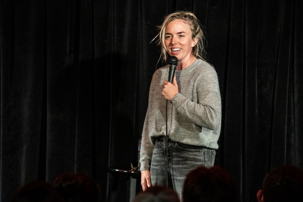 A woman holding a mic smiling wearing a grey sweater