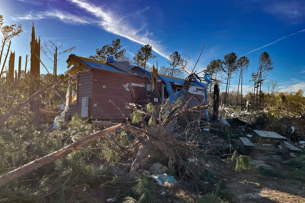 A browinsh red home sits in the middle of a wooded area with broken trees surrounding it.