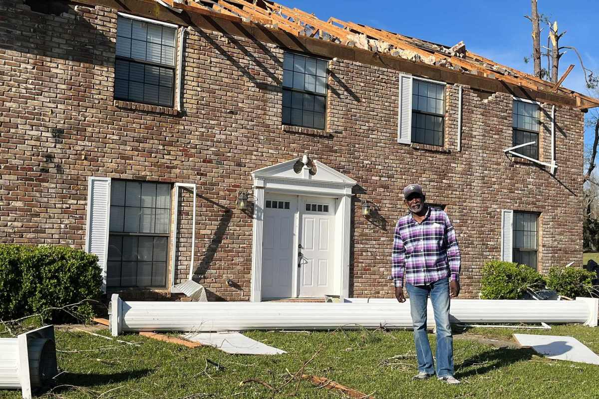 A man in a plaid shirt stands in front of a brown brick home with the roof exposed and a white column laying behind him on the ground.