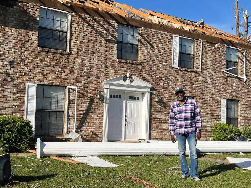 A man in a plaid shirt stands in front of a brown brick home with the roof exposed and a white column laying behind him on the ground.