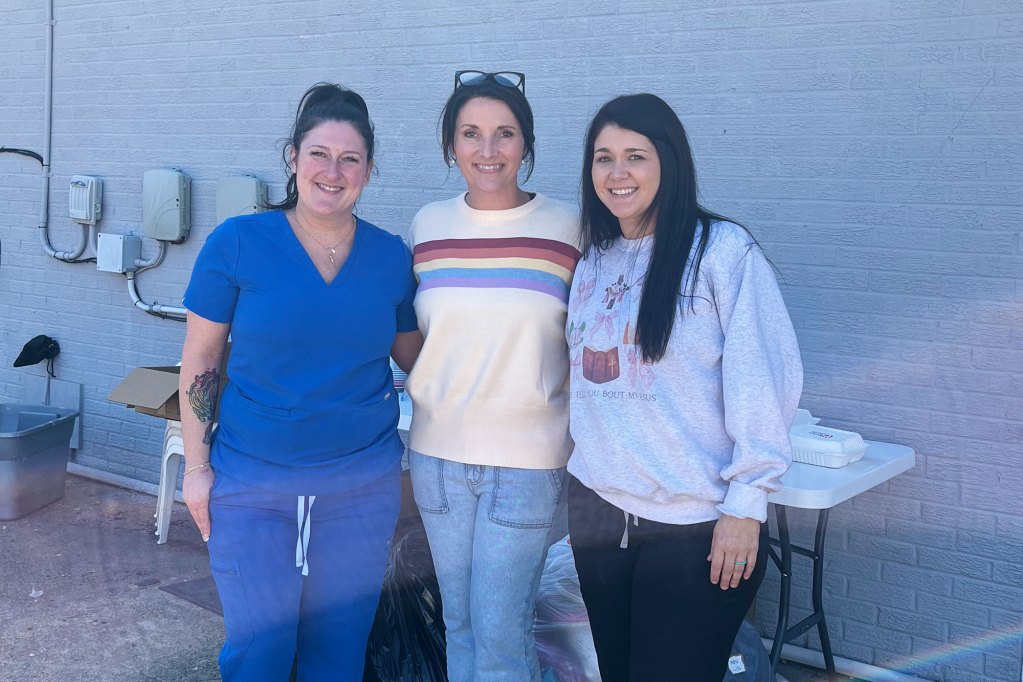 Three women stand together for a photo in front a grey brick wall.