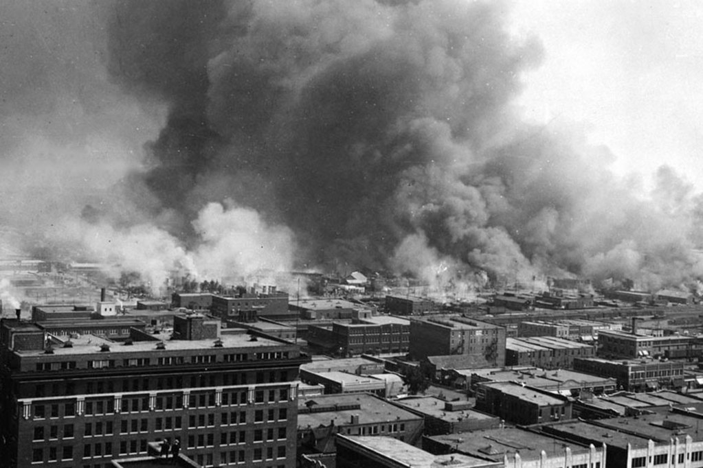 A black and white photo of buildings in the foreground and a cloud of smoke in the background.