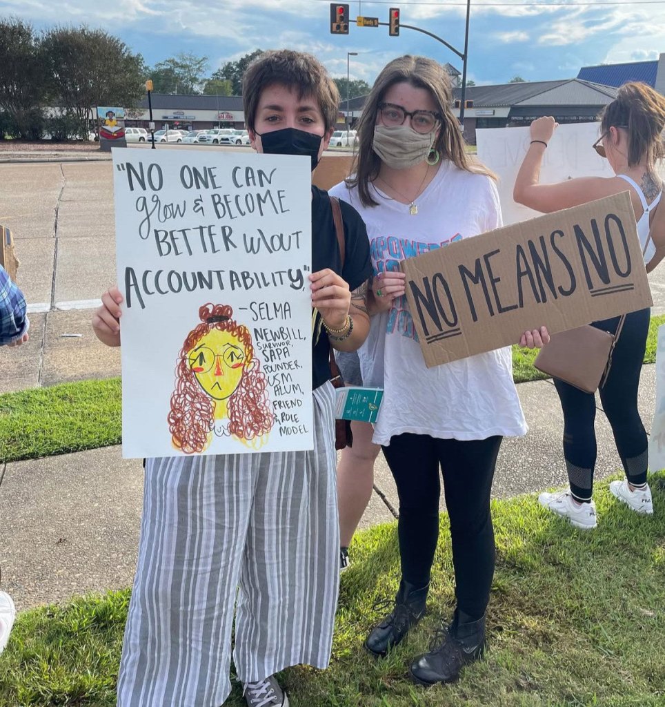 Two women holding protest signs standing in grass with a street behind them
