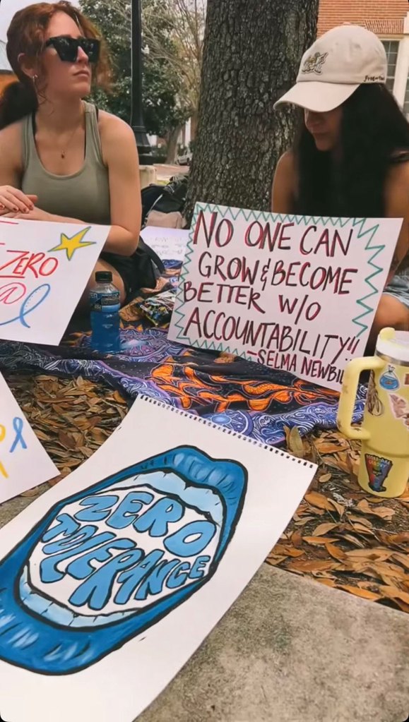 Two women sitting on the ground holding protest signs with a zero tolerance sign in white and blue positioned in front of them