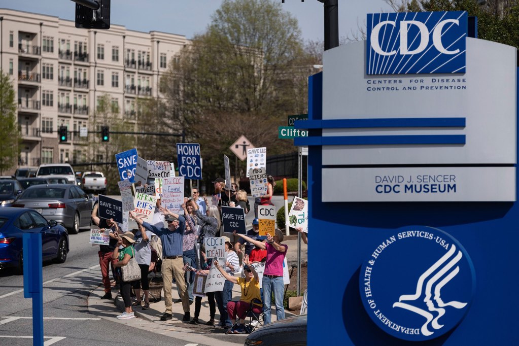People protesting right next to a blue and grey CDC sign.