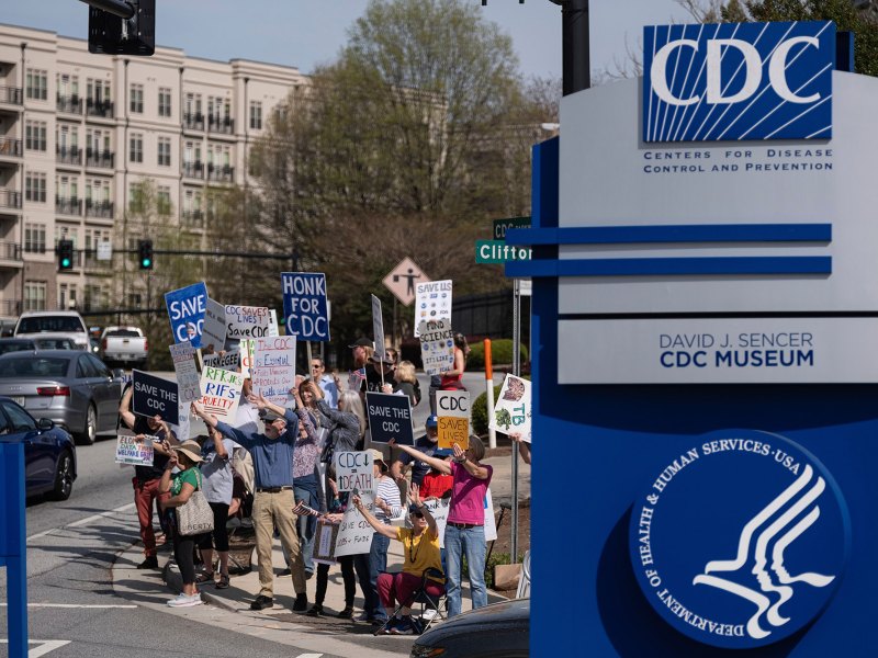 People protesting right next to a blue and grey CDC sign.