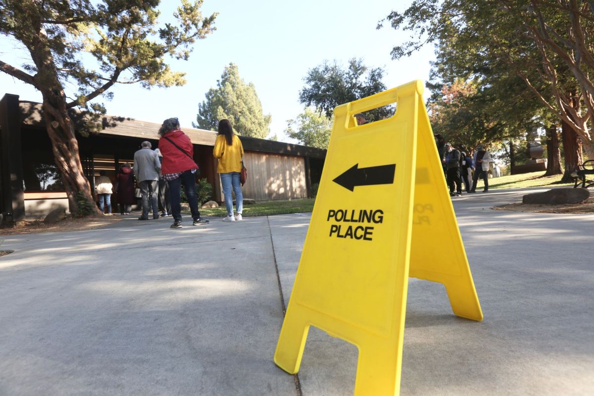People line outside of a building past a bright yellow sign that reads Polling Place and points toward the building