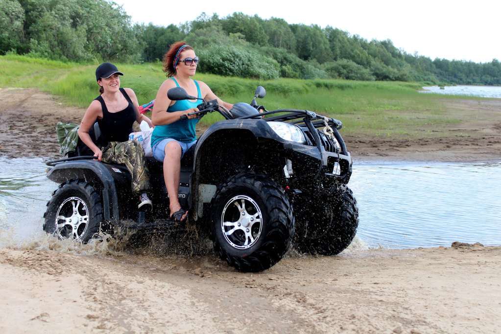 Two women riding on a black atv moving through water and dirt with green land in the background