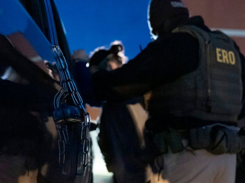 A rear view showing a police agent in a jacket labeled ERO arresting someone against a car