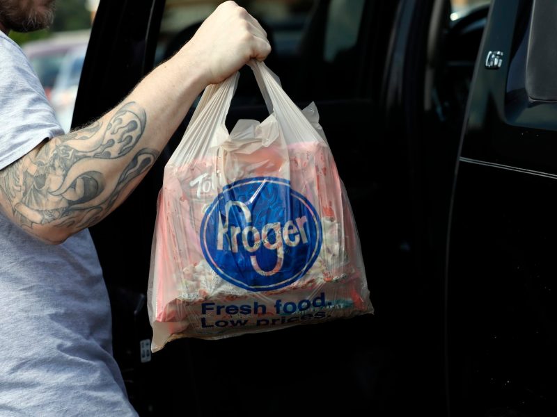 a photo of someone’s tattooed arm as they lift a grocery bag with the word “Kroger” on it into a car