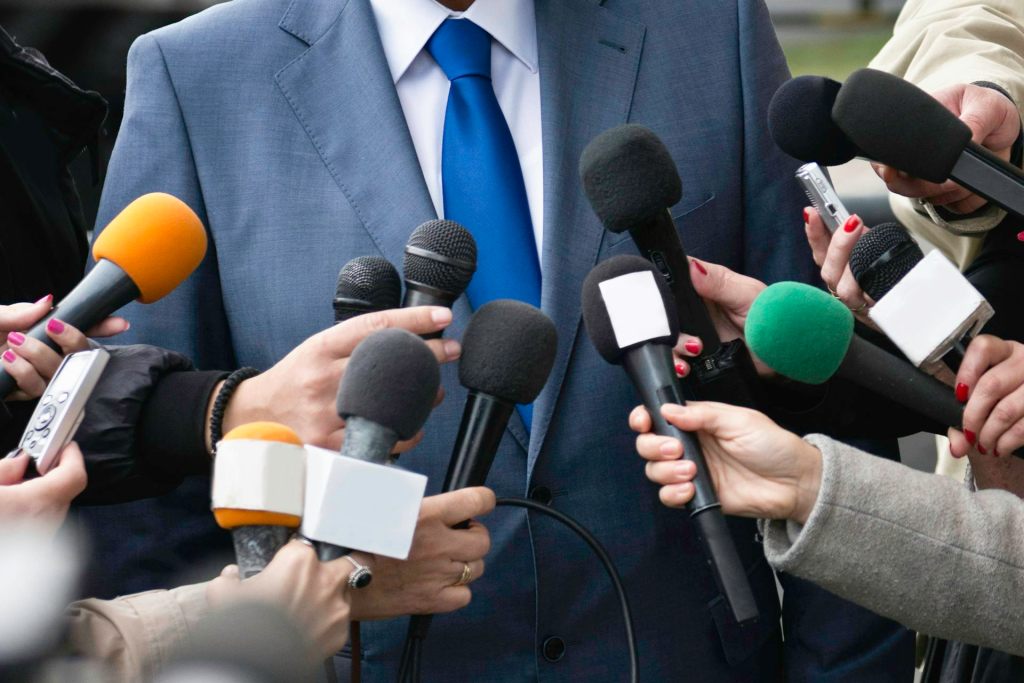 About a dozen microphones are held out toward a man in a suit and tie, whose face is outside the frame of the photo