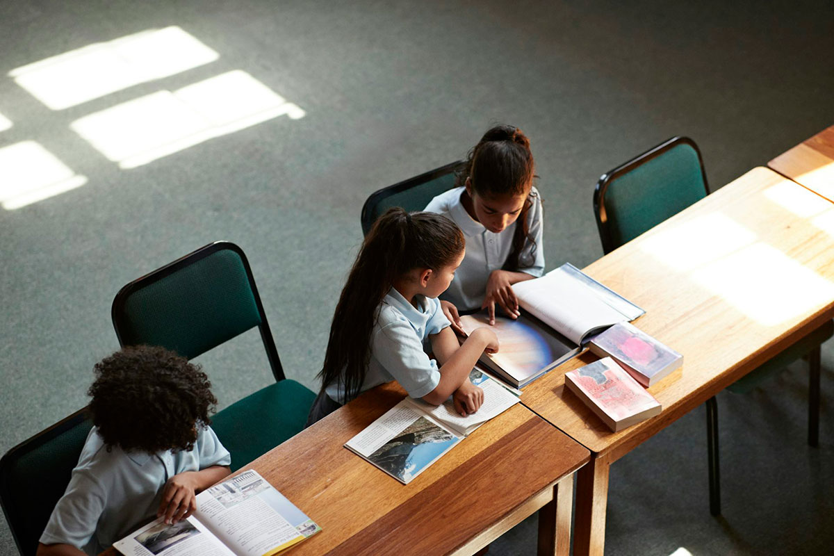 Three students sit at a desk while they read books
