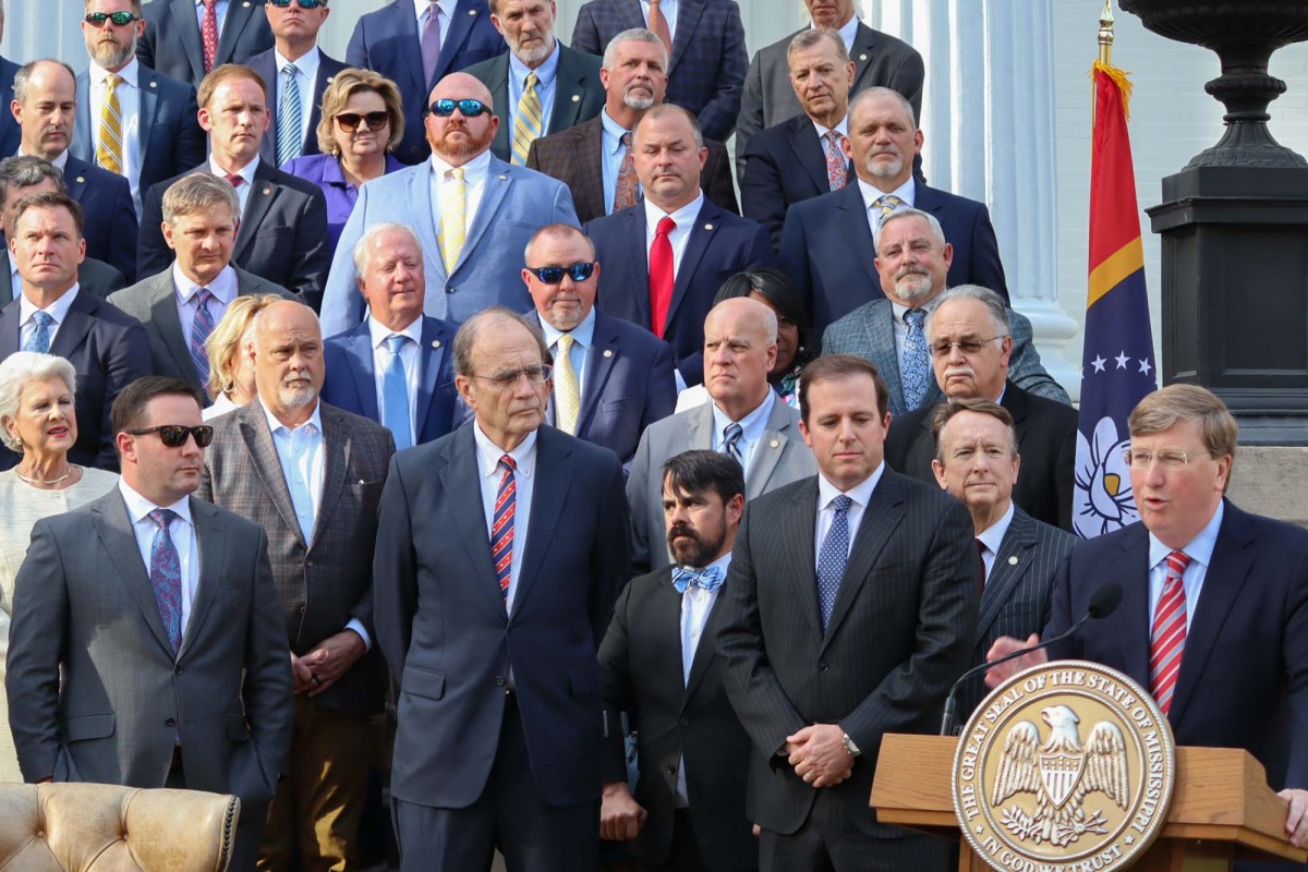 a photo of tate reeves speaking at a lectern with delbert hosemann and state lawmakers gathered behind him