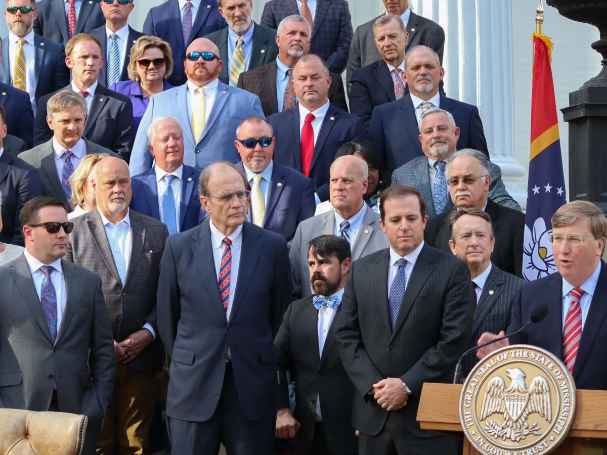 a photo of tate reeves speaking at a lectern with delbert hosemann and state lawmakers gathered behind him
