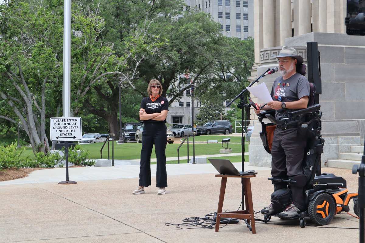 A woman in all black stands and looks at a man speaking wearing grey