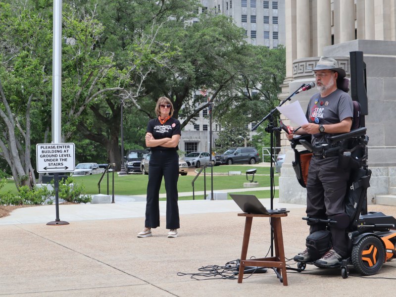 A woman in all black stands and looks at a man speaking wearing grey