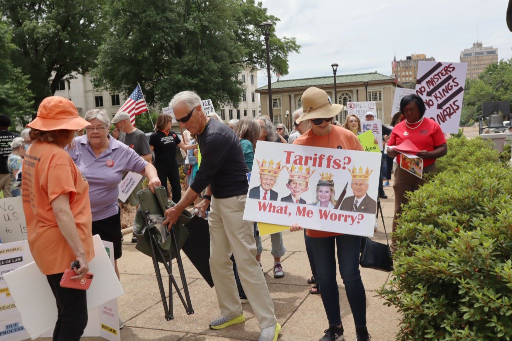 a photo shows protestors, including a woman holding a sign that says "Tariffs? What, me worry?" with images of of Donald Trump, Alfred E. Neuman, Cindy Hyde-Smith and Roger Wicker all weaaring crowns