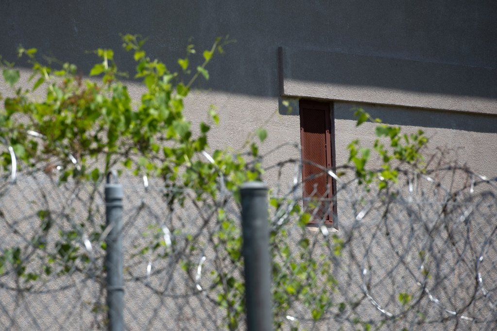A close up photo of a narrow window covered by a brown-red metal grate, with barbed wire and leaves in the foreground.
