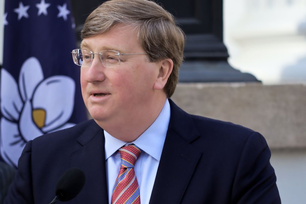 Governor Tate Reeves wearing a navy suit jacket, powder blue shirt, and red striped tie speaks at a podium