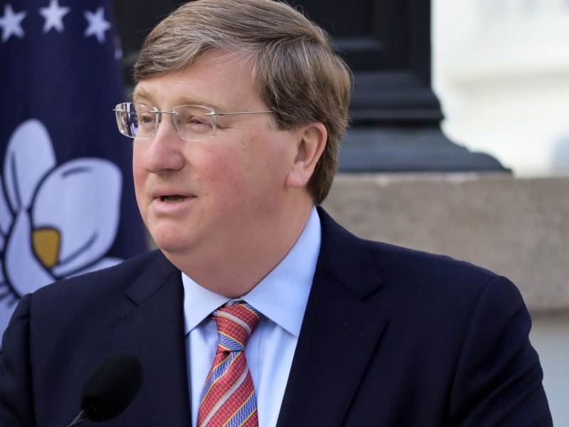 Governor Tate Reeves wearing a navy suit jacket, powder blue shirt, and red striped tie speaks at a podium