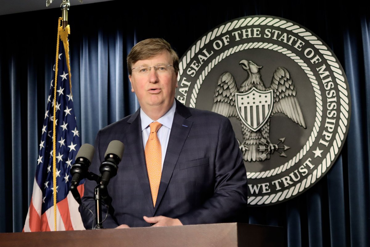 Governor Tate Reeves speaking at a press conference with an American flag next to him and a State of Mississippi official seal behind him
