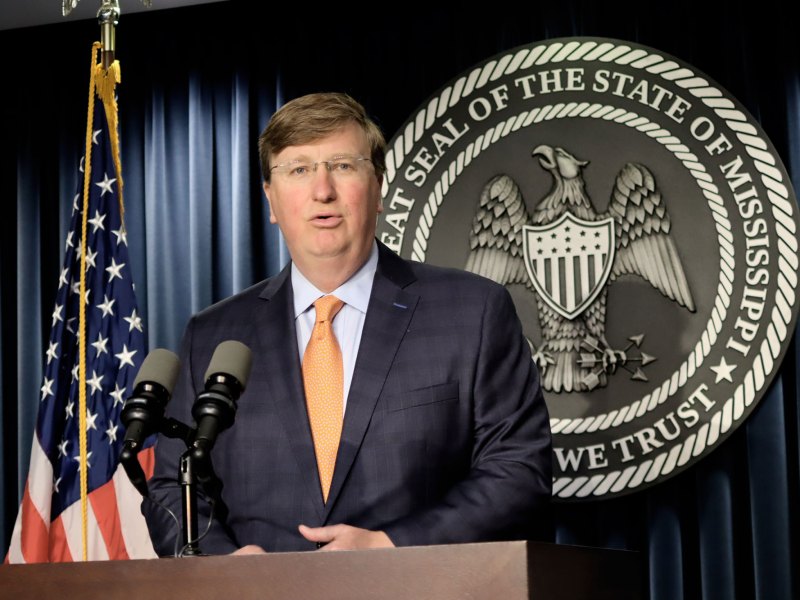 Governor Tate Reeves speaking at a press conference with an American flag next to him and a State of Mississippi official seal behind him