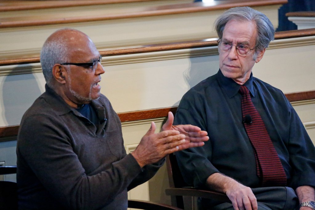 Two men speak together while seated on stage