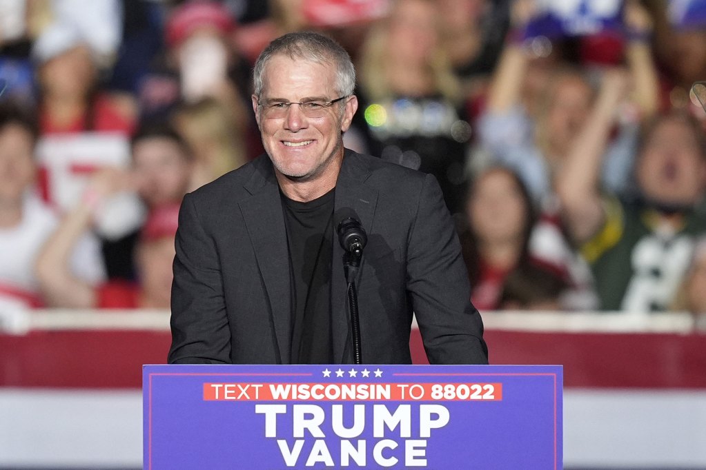 Brett Favre in a dark suit and glasses smiles at a podium labeled TRUMP VANCE