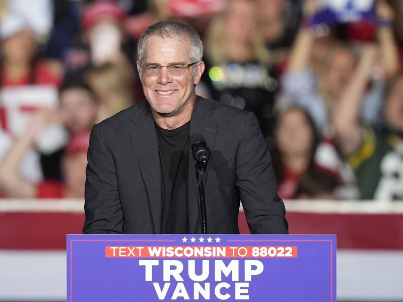 Brett Favre in a dark suit and glasses smiles at a podium labeled TRUMP VANCE